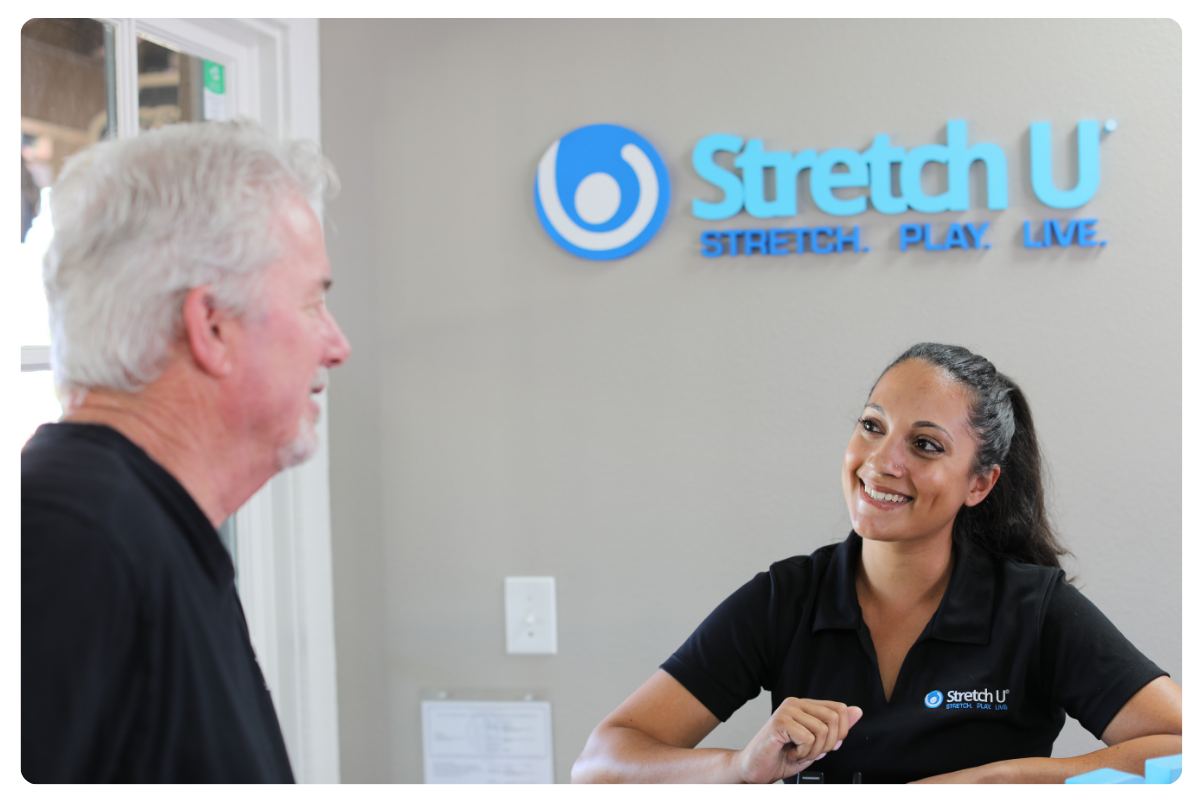 A friendly Stretch U team member warmly welcomes a senior male client at the reception desk of our Greeley, CO studio, with the "Stretch. Play. Live." logo visible.