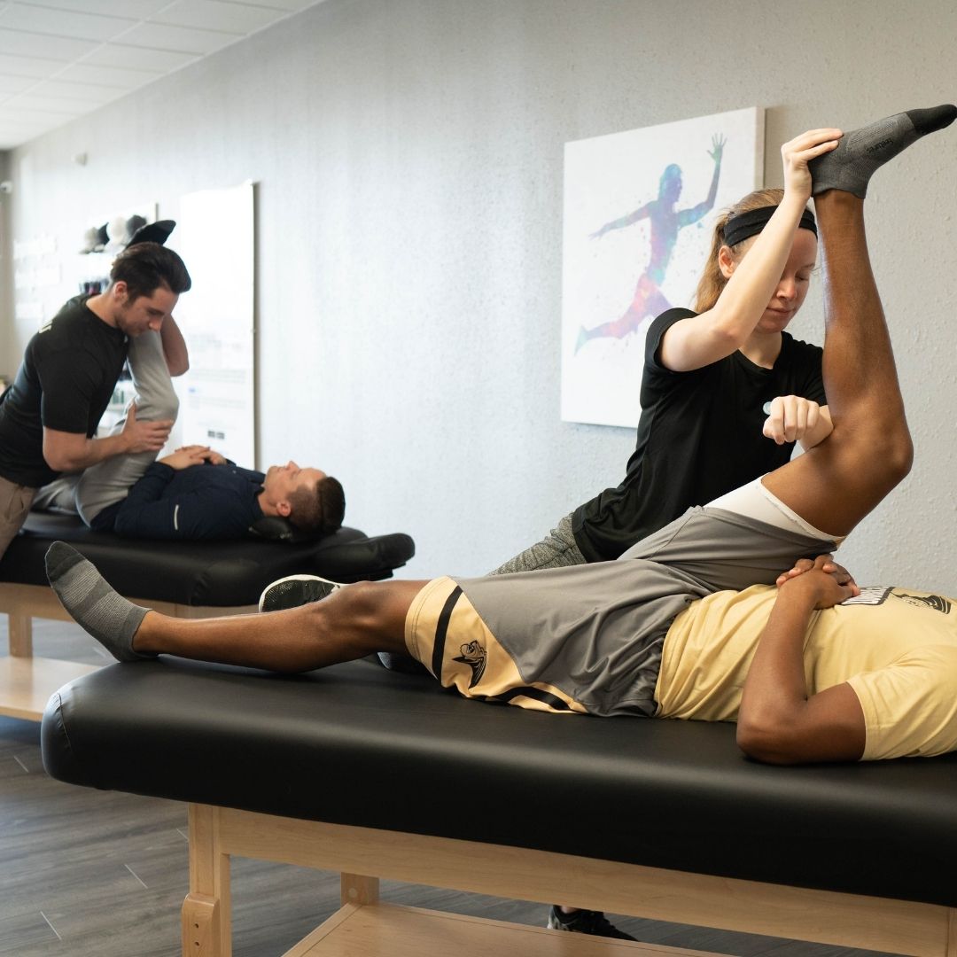 Two male clients receive assisted leg stretches from two therapists simultaneously in a professional stretching studio.