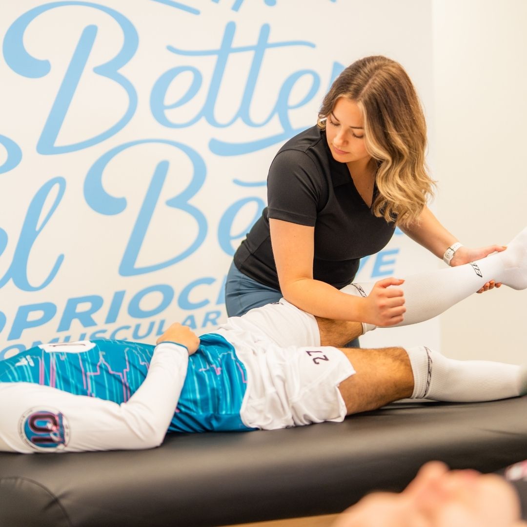 A female therapist performs a deep assisted leg stretch on an athlete in a soccer jersey.