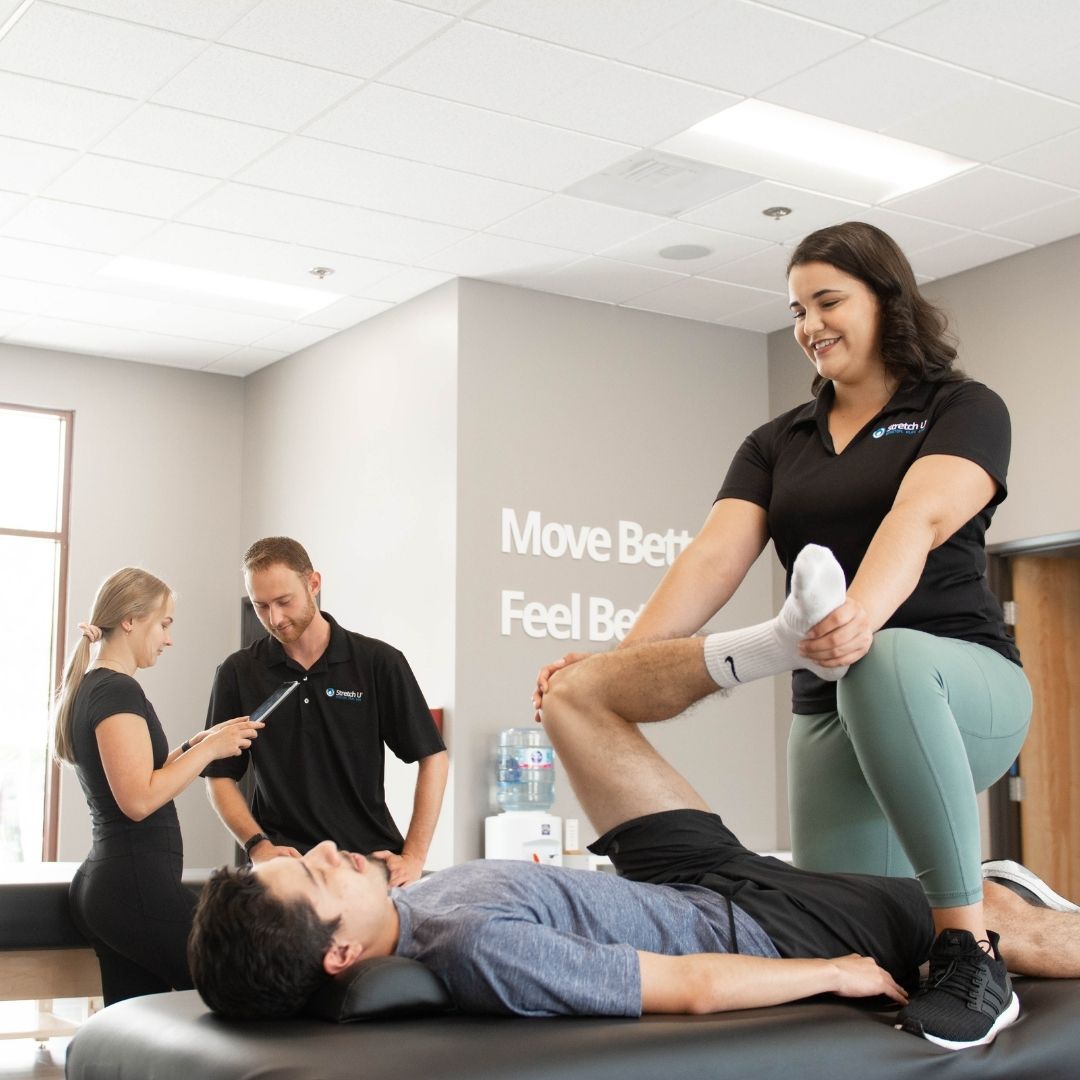 A female therapist assists a male client with a leg stretch while two other staff members observe or consult in the background of the studio.