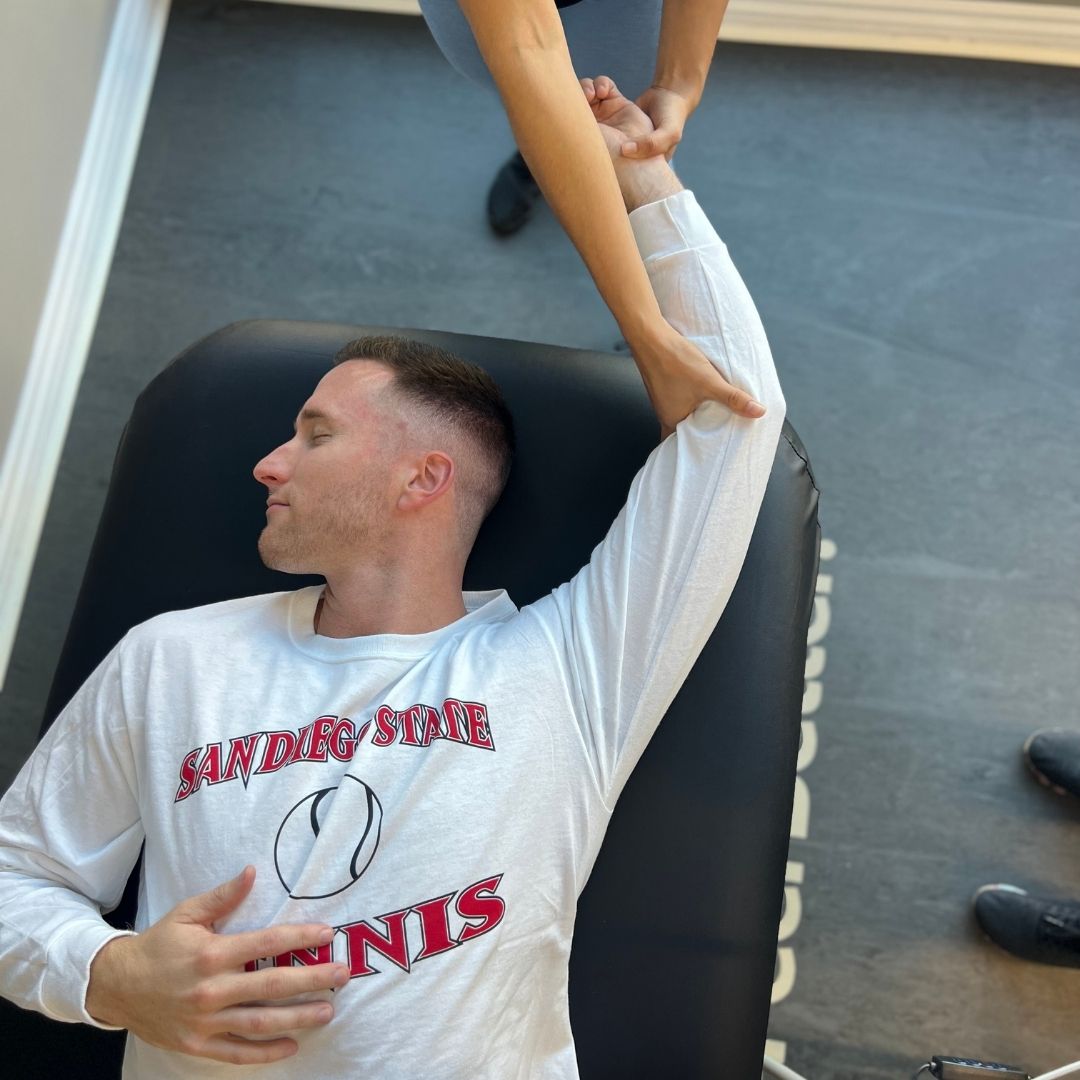 A male client wearing an athletic long-sleeve shirt receives an assisted arm and shoulder stretch from a therapist.