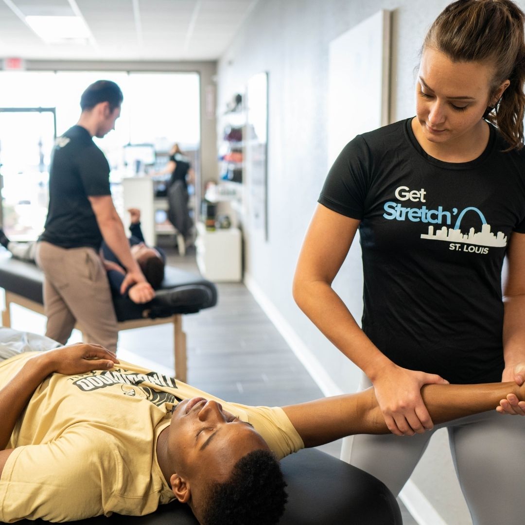 A female therapist in the foreground performing an assisted arm/shoulder stretch on a male client lying on a table in a brightly lit studio.