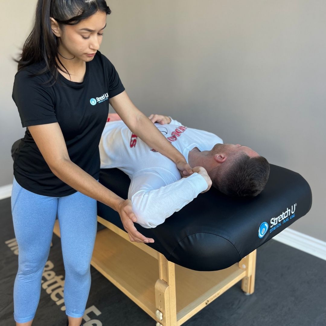 A female therapist performs an assisted shoulder and neck stretch on a male client on a portable table, demonstrating an on-site stretch session.