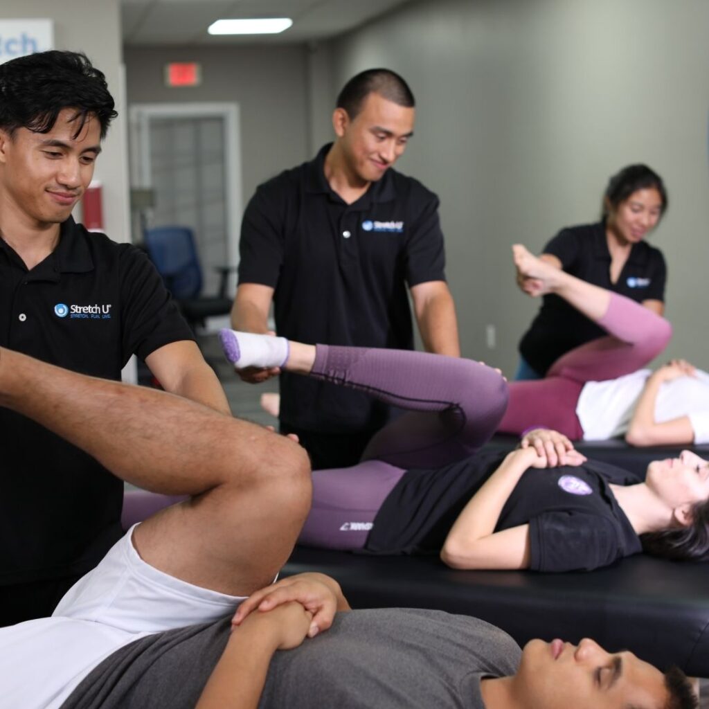 Two male therapists perform assisted leg stretches on two clients side-by-side in a multi-client setting, highlighting group or corporate stretching.