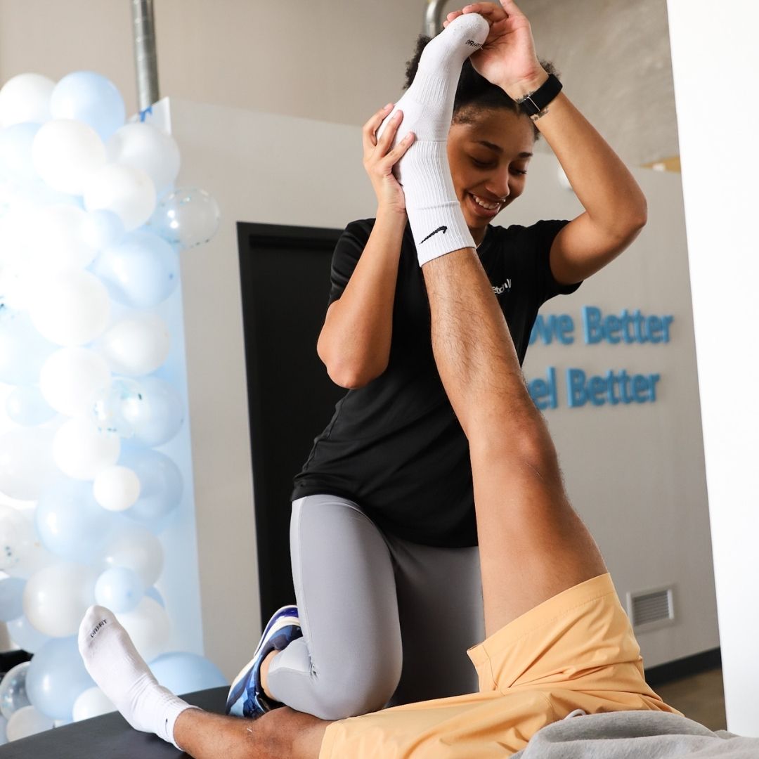 A smiling female therapist performs a high leg stretch on a male client at an event or brightly decorated pop-up wellness station.