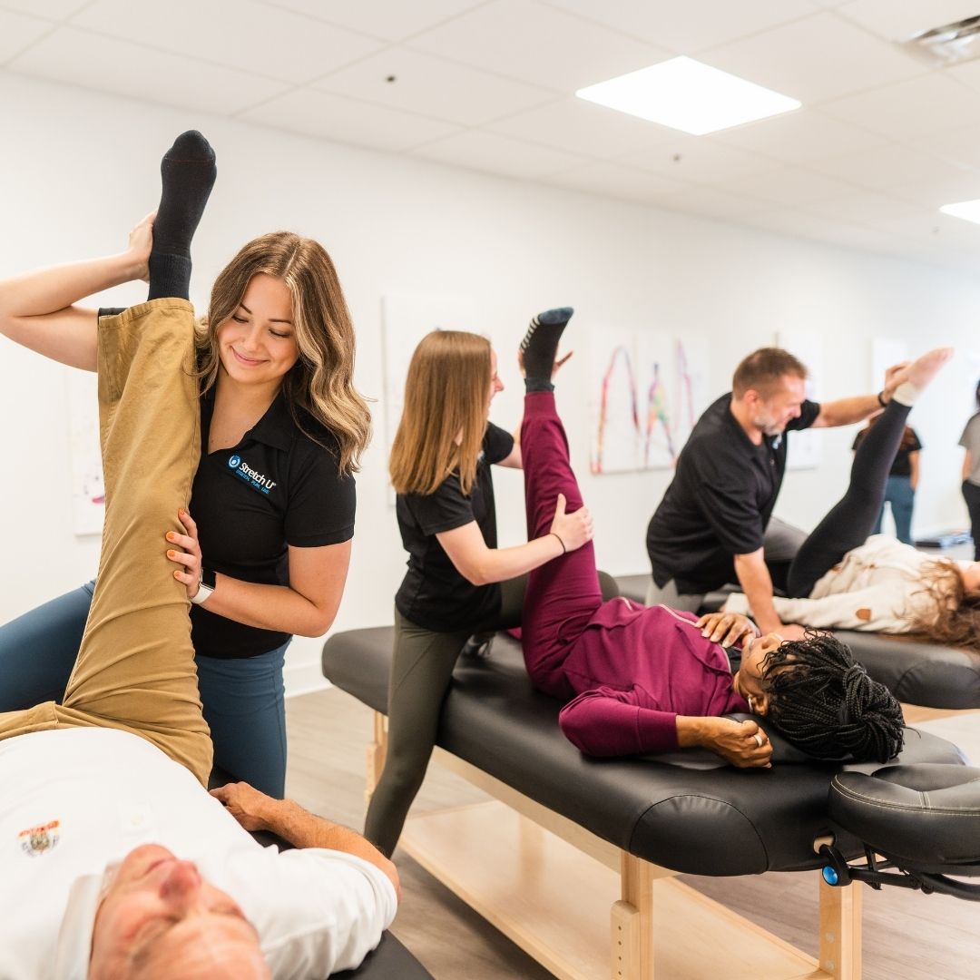 A wide shot showing multiple therapists performing assisted leg stretches on clients lying on tables, demonstrating a busy, professional group stretching environment.