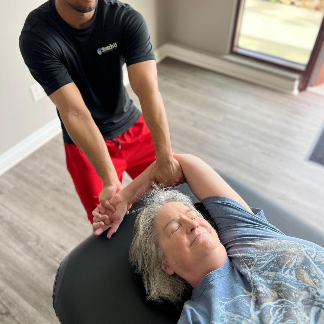 A male therapist performs an assisted arm and shoulder stretch on an older female client who appears relaxed on the stretching table.
