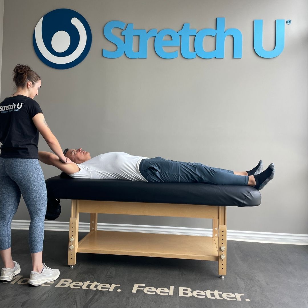 A female therapist performs an assisted arm stretch on a male client lying supine on a stretching table in a professional studio setting.